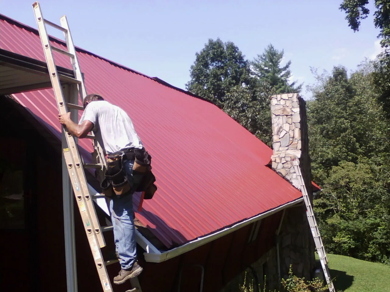 Roofer on a ladder beside a red metal roof and stone chimney — Soffit & Fascia Repair by Roofing Brothers in Rocky Hill